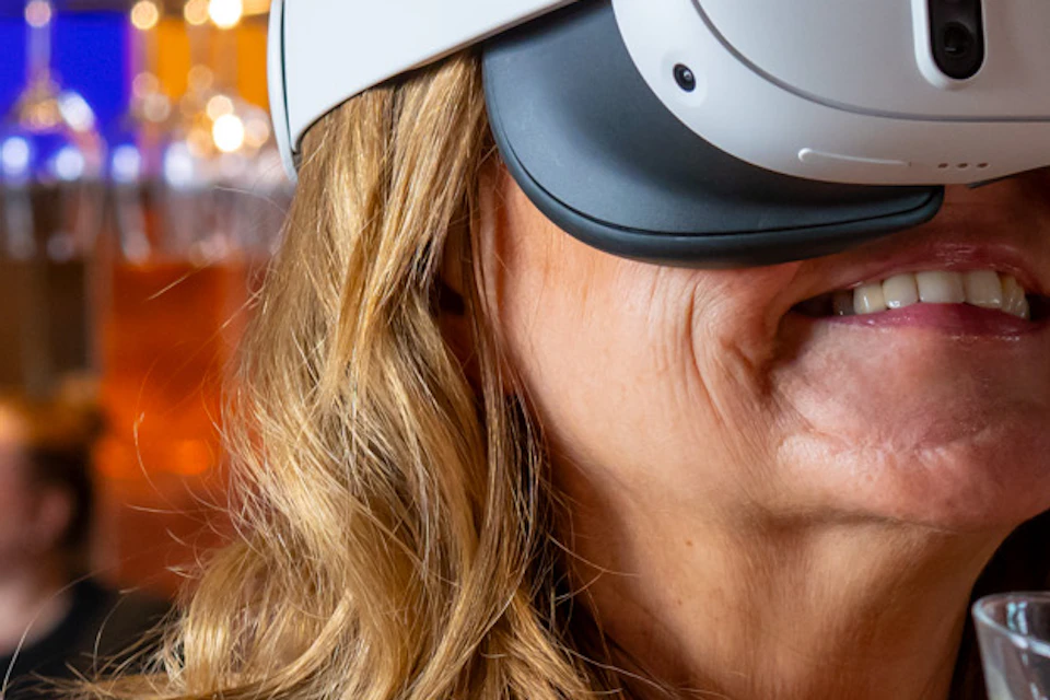 A woman wearing a VR headset holds a small glass of liquid in a bar setting, with shelves of bottles in the background.