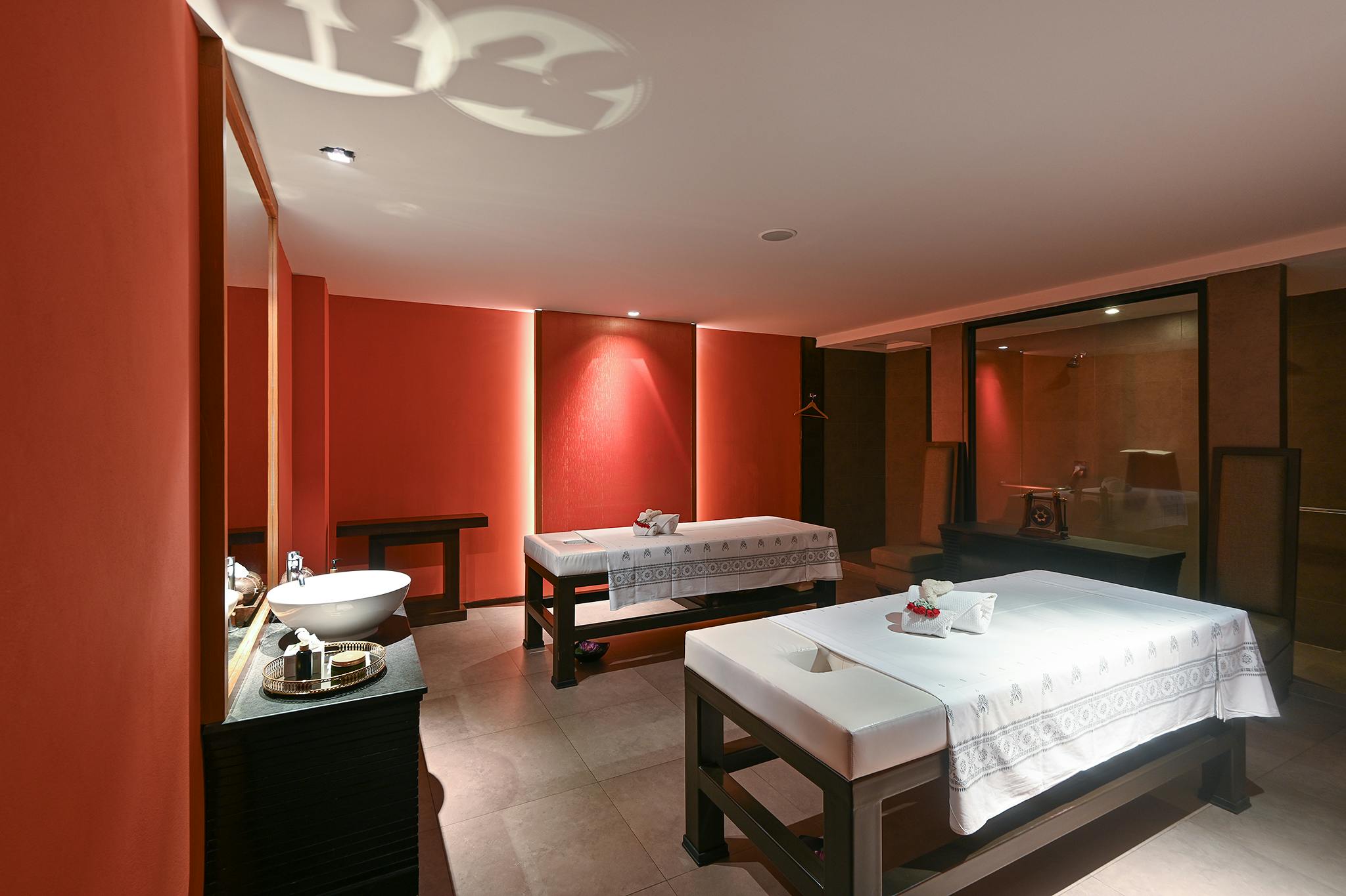 A spa treatment room with two massage tables, red walls, a sink, and neatly arranged towels in a calming ambiance.