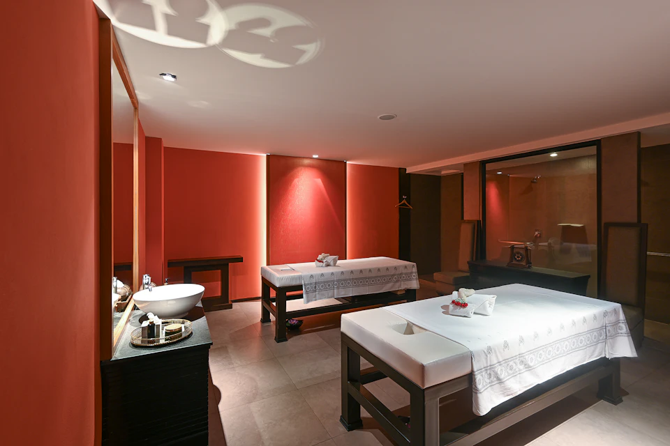 A spa room with two massage tables, a sink, and warm lighting, featuring red walls and a modern, serene design.