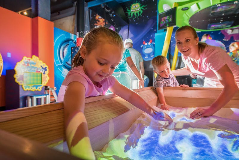 Two children and a woman play with interactive sand in a brightly-lit, space-themed exhibit.