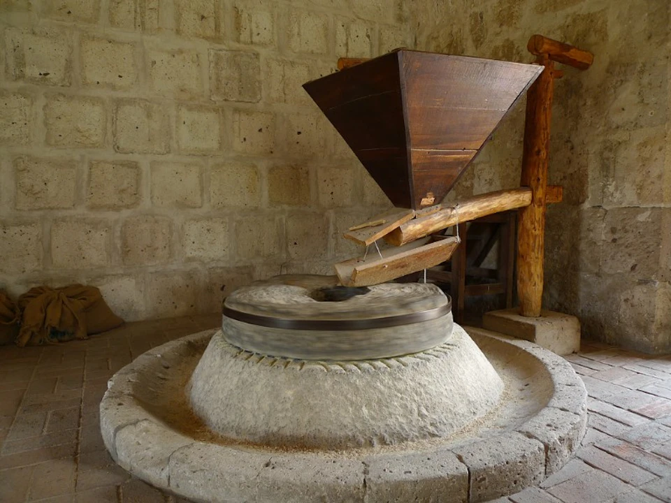An old-fashioned stone mill with a wooden hopper stands in a rustic room with stone walls and wooden beams.