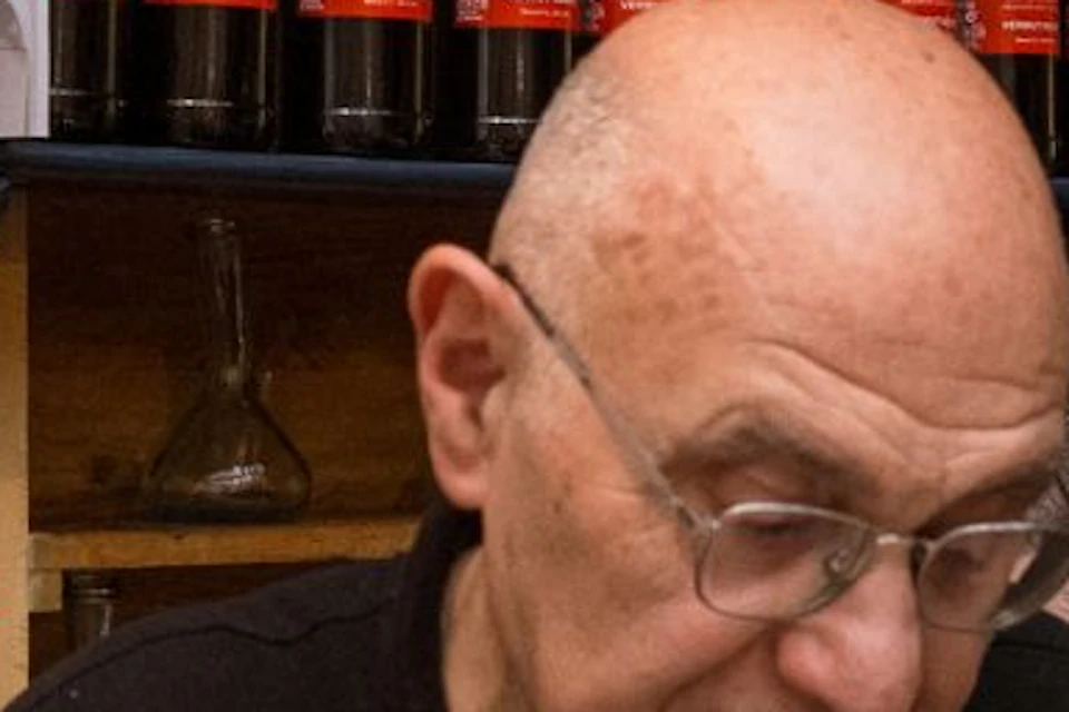 A man in a black shirt is reading a newspaper behind a counter in a shop with shelves of bottled drinks on display.