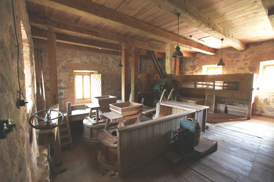 Old wooden interior of a mill with traditional grinding equipment, stone walls, exposed beams, and dim lighting from small windows.