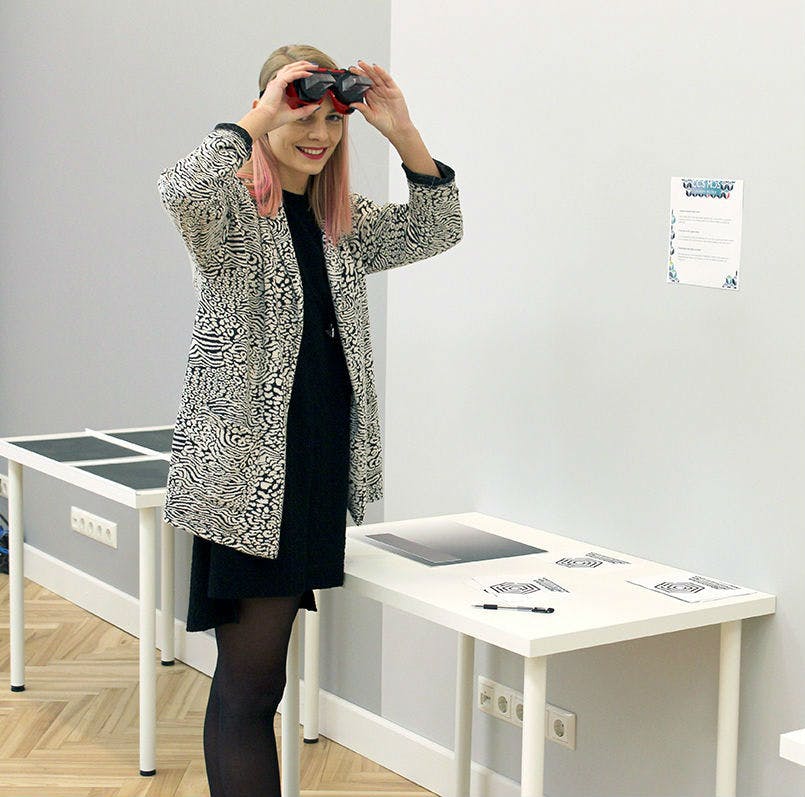 站在桌子旁边的人，穿着带图案的外套，微笑着，将红色和黑色的虚拟现实耳机戴在眼睛上。