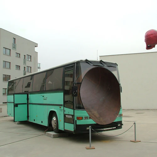 En turkos turnébuss med en stor högtalarliknande struktur på baksidan parkerad på en innergård med moderna byggnader och en röd skalle-skulptur.
