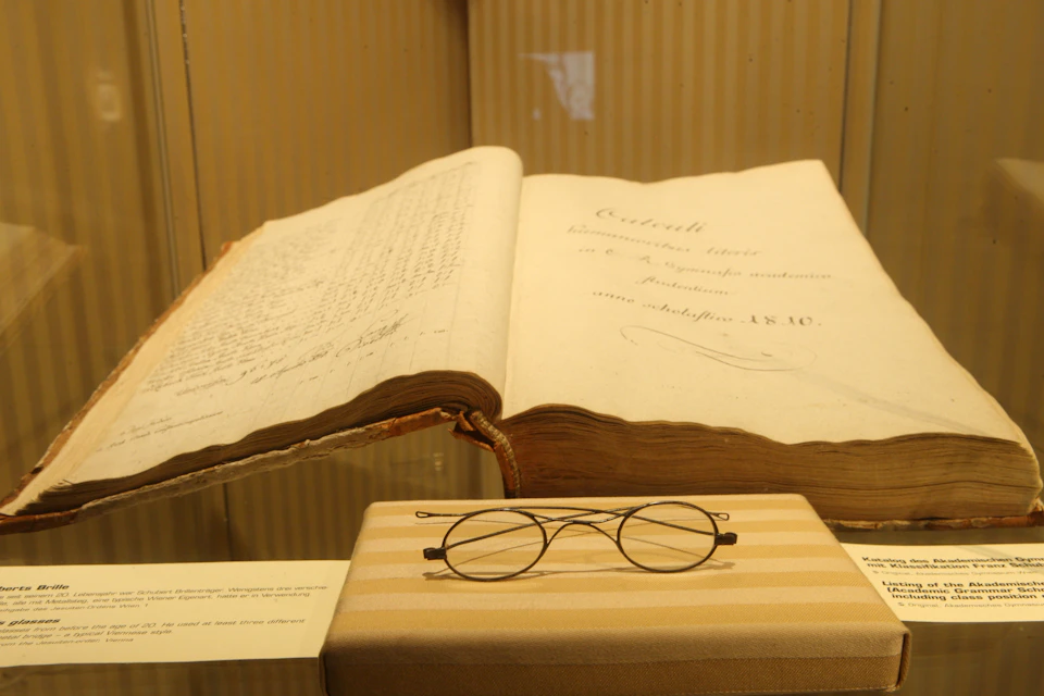 An open antique book is displayed in a glass case, with round wire-rimmed glasses resting on a striped cloth below it.