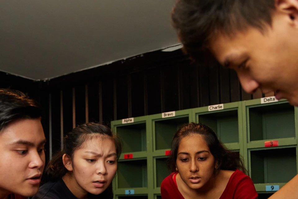 Four people gather around a table, examining an object, with shelves labeled with names in the background.