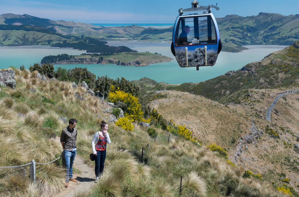 Two people walking on a hillside path with a cable car passing overhead, overlooking a scenic landscape with hills and water.