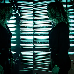 En person med lockigt hår står framför spegelväggar i ett mörkt rum med blågröna neonljusreflektioner, vilket skapar flera reflektioner.