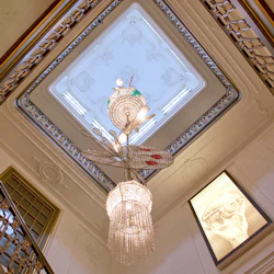 Tittar upp mot ett utsmyckat tak med en stor kristallkrona, invecklade räcken och en inramad arkitektonisk ritning.