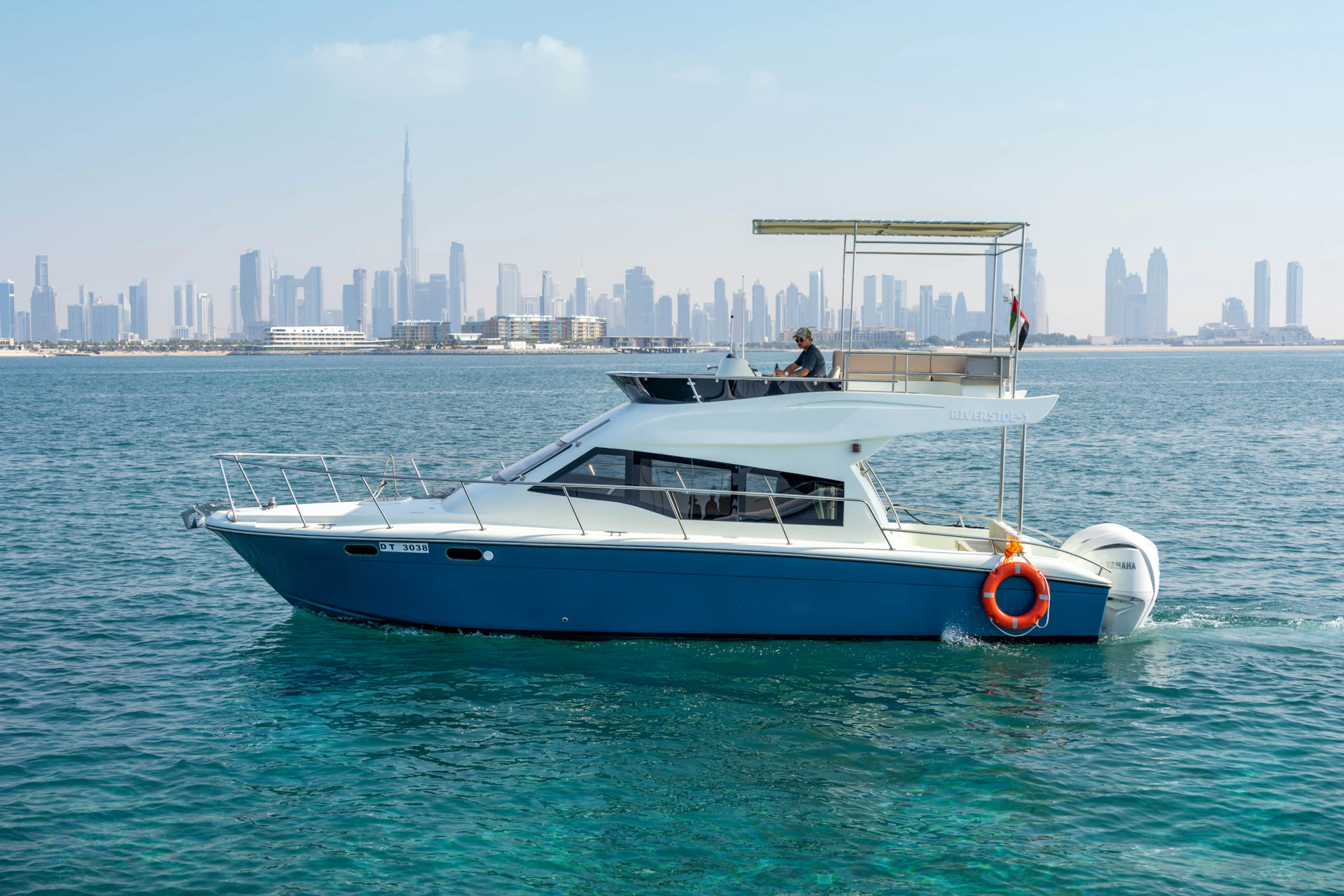A motorboat with a blue hull and white cabin on the water, with a city skyline and the Burj Khalifa visible in the background.