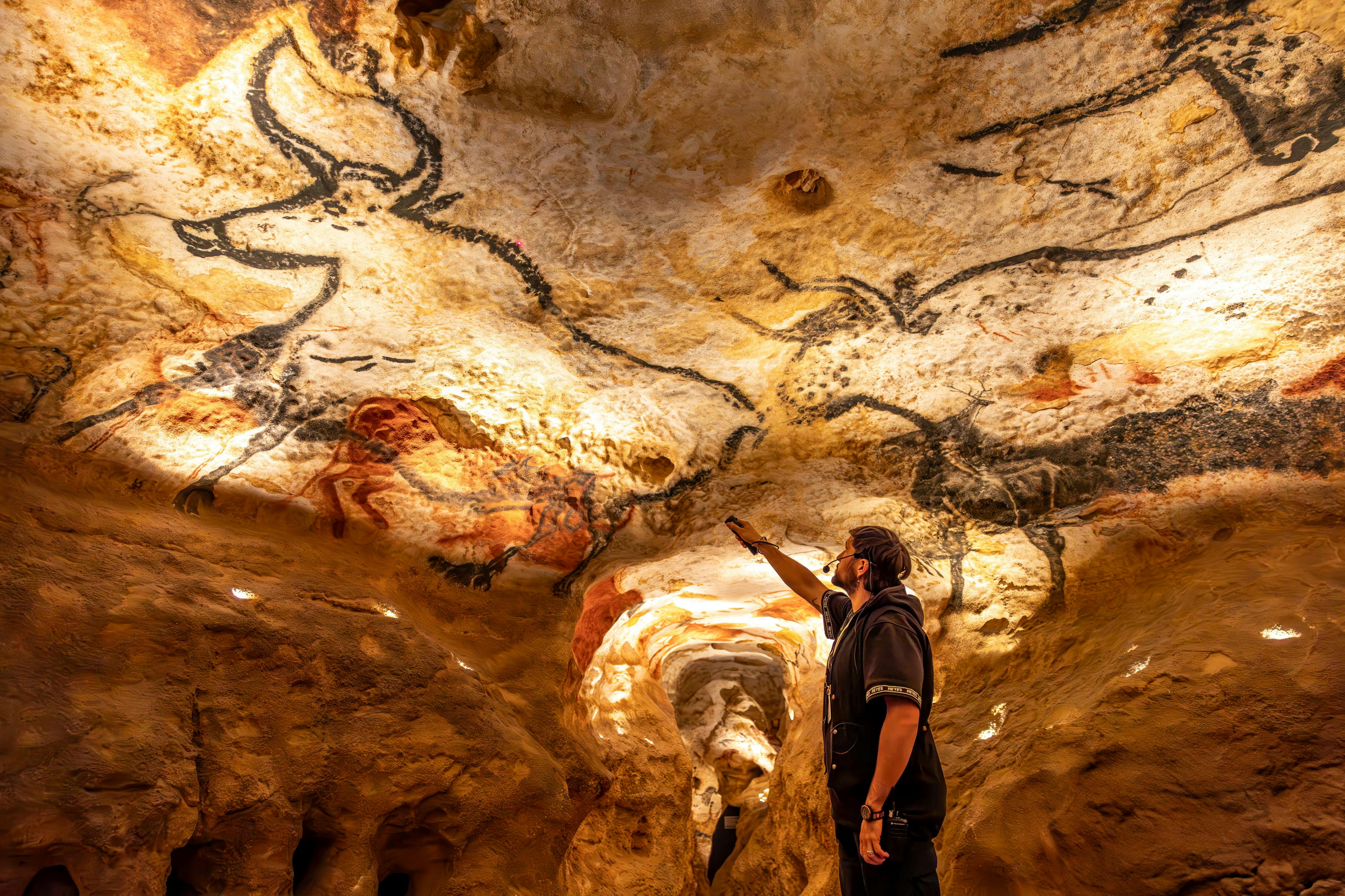 A person pointing at prehistoric cave paintings of animals on a brightly lit cave ceiling.