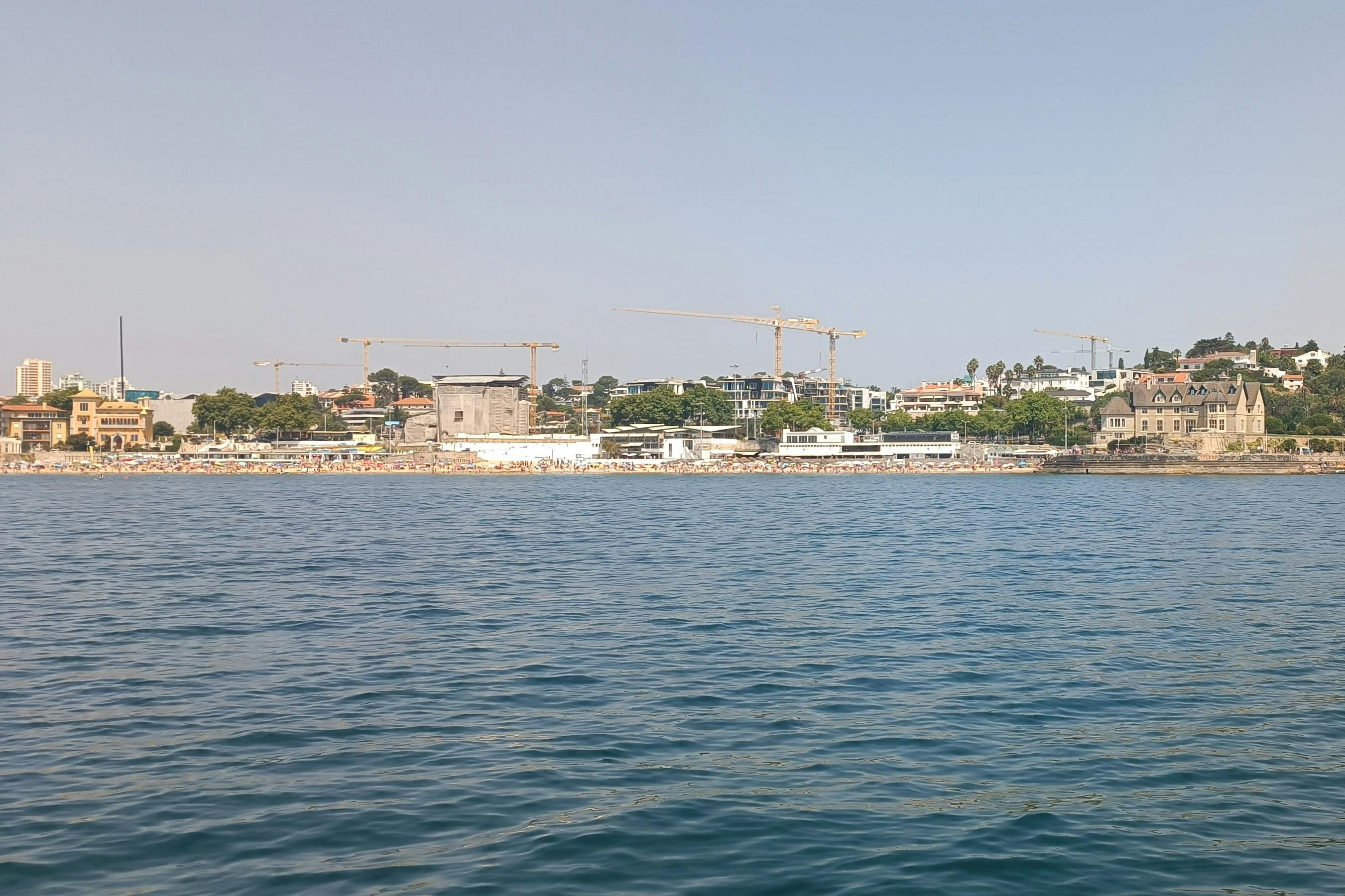 Παράκτια σκηνή κατασκευής με γερανούς, κτίρια και πράσινα δέντρα στο παρασκήνιο, όπως φαίνεται από το νερό.