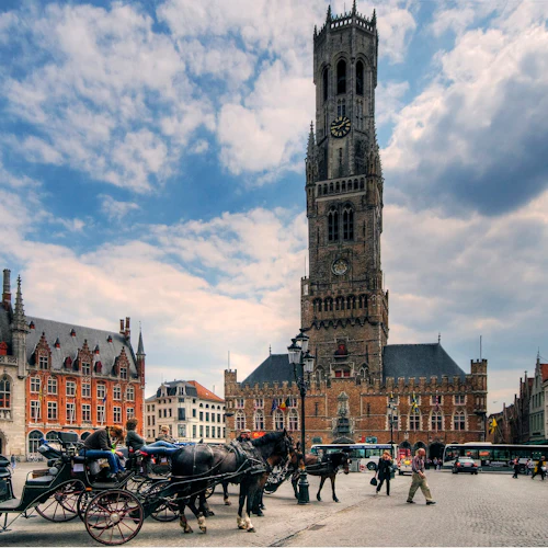 Hästar och vagnar på ett kullerstensbelagt torg med ett stort medeltida klocktorn och historiska byggnader i bakgrunden.