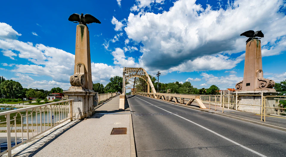 An empty road bridge with a large bird statue pillar, under a blue sky with scattered clouds. Trees are visible in the background.
