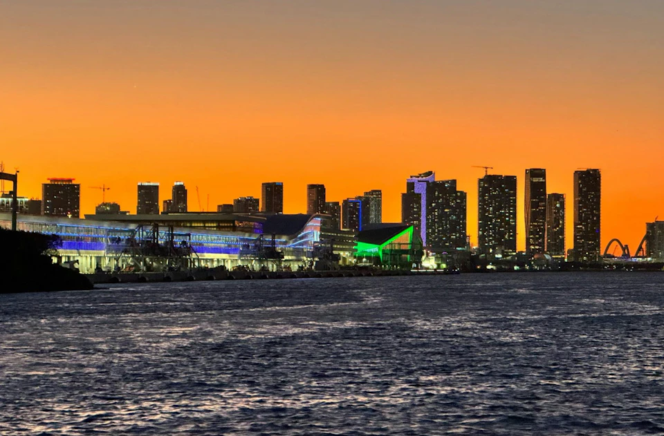City skyline at sunset with illuminated buildings, including a green-lit structure, viewed from across the water.