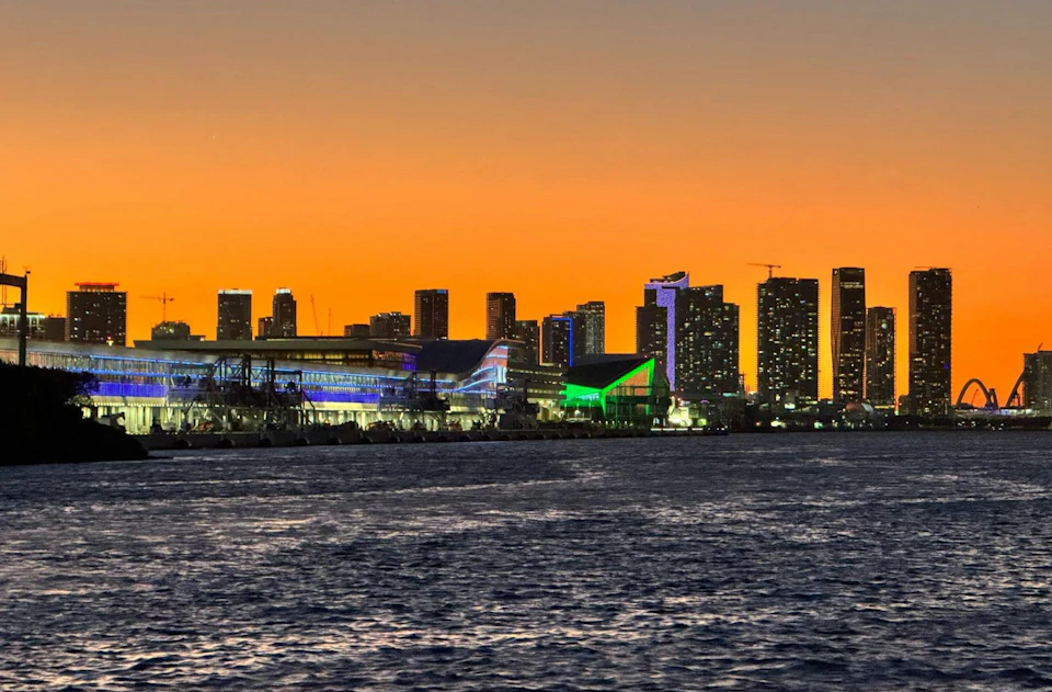 City skyline at sunset with illuminated buildings, including a green-lit structure, viewed from across the water.