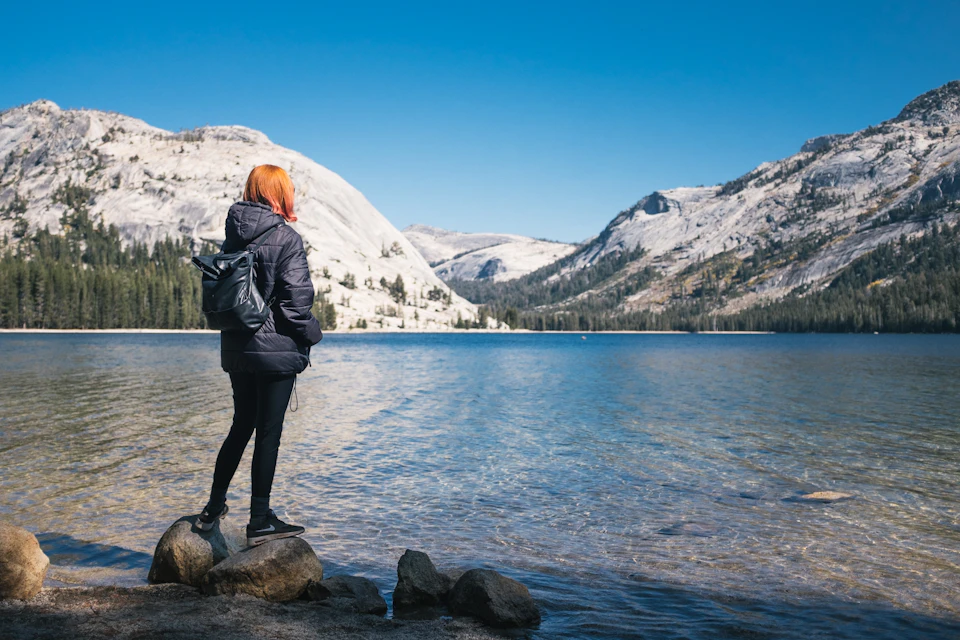 A person with red hair and a backpack stands on rocks at the edge of a lake, surrounded by mountains under a clear blue sky.