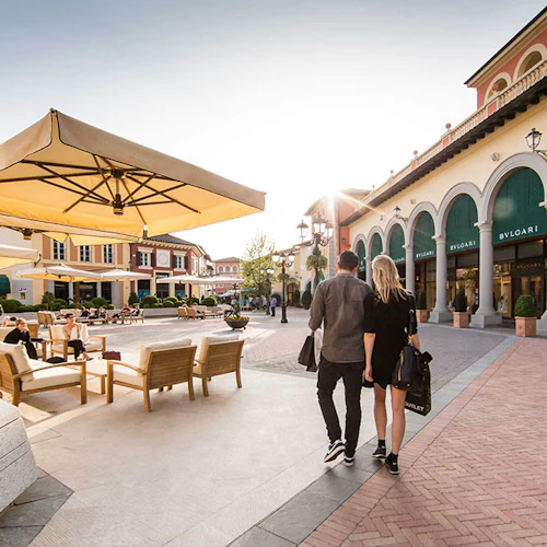 Utomhus shoppingtorg med välvda skyltfönster, stora paraplyer, sittgrupp och ett par som går medan de håller i shoppingväskor.