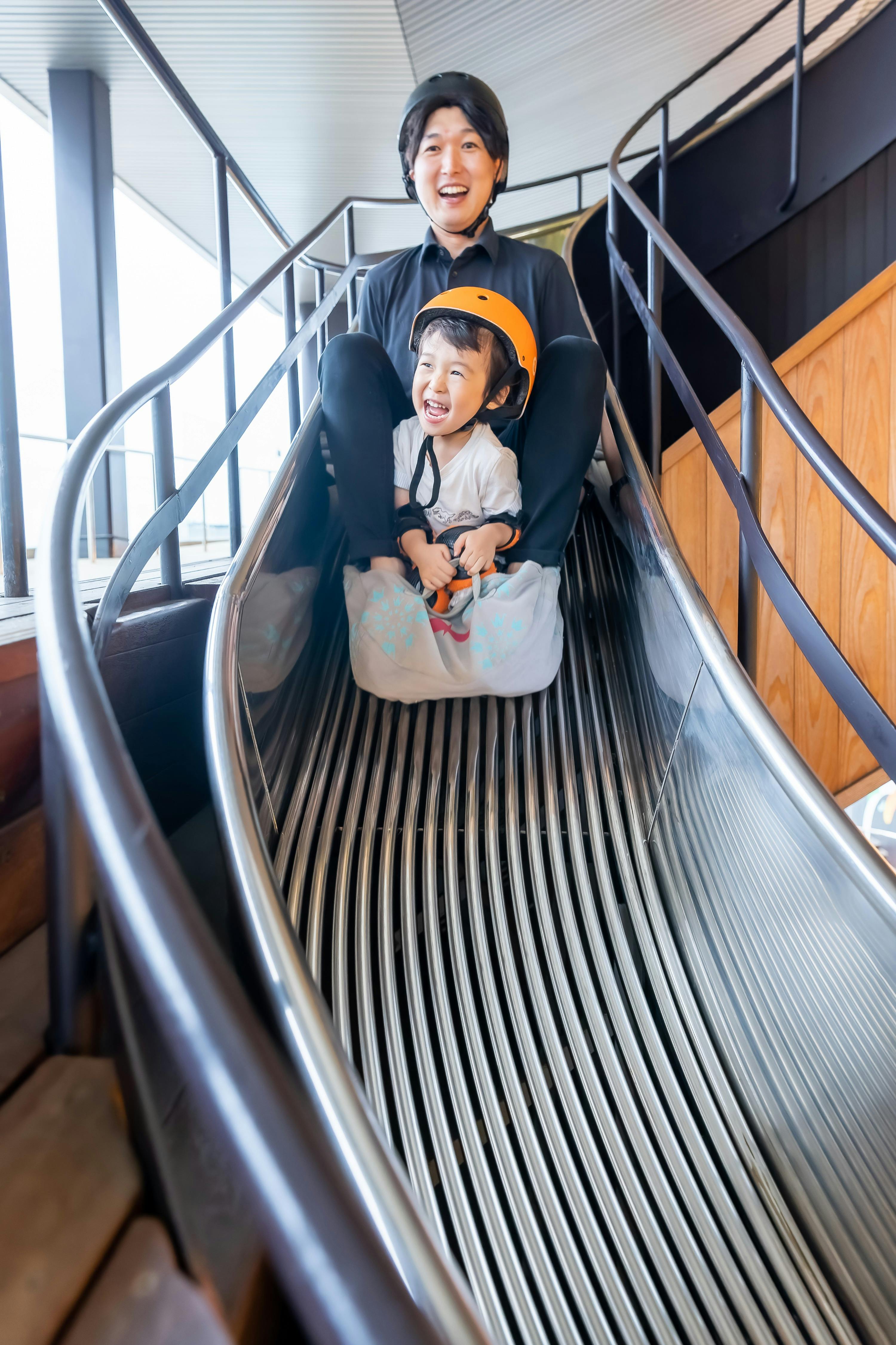 An adult and a child wearing helmets sliding down a metal slide indoors, both smiling and looking excited.