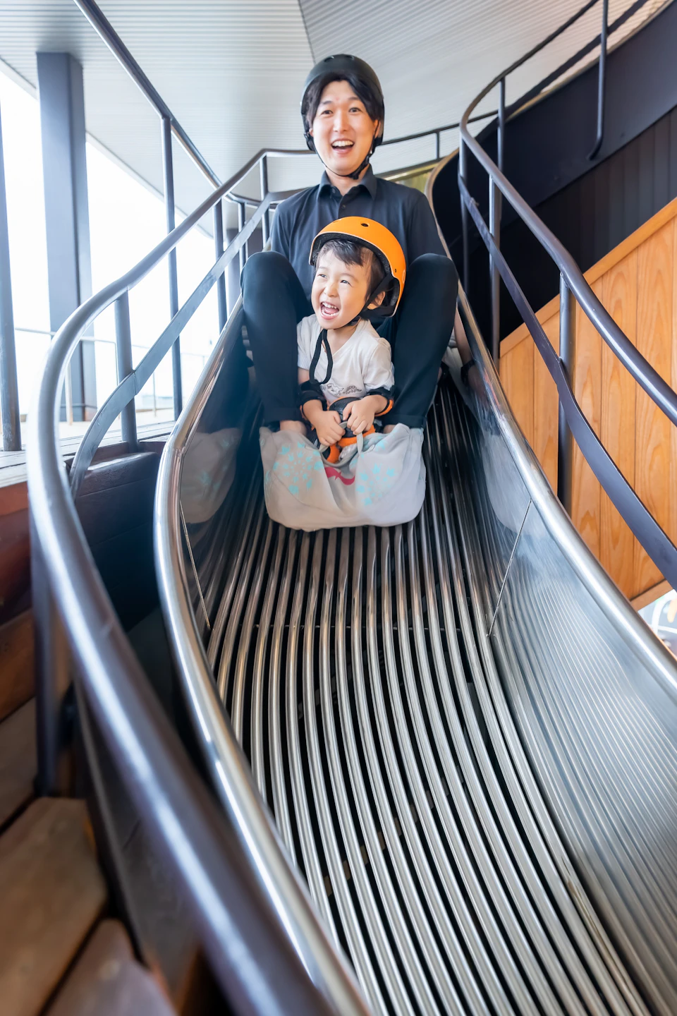 An adult and a child wearing helmets slide down a metal slide together indoors, smiling and enjoying the ride.