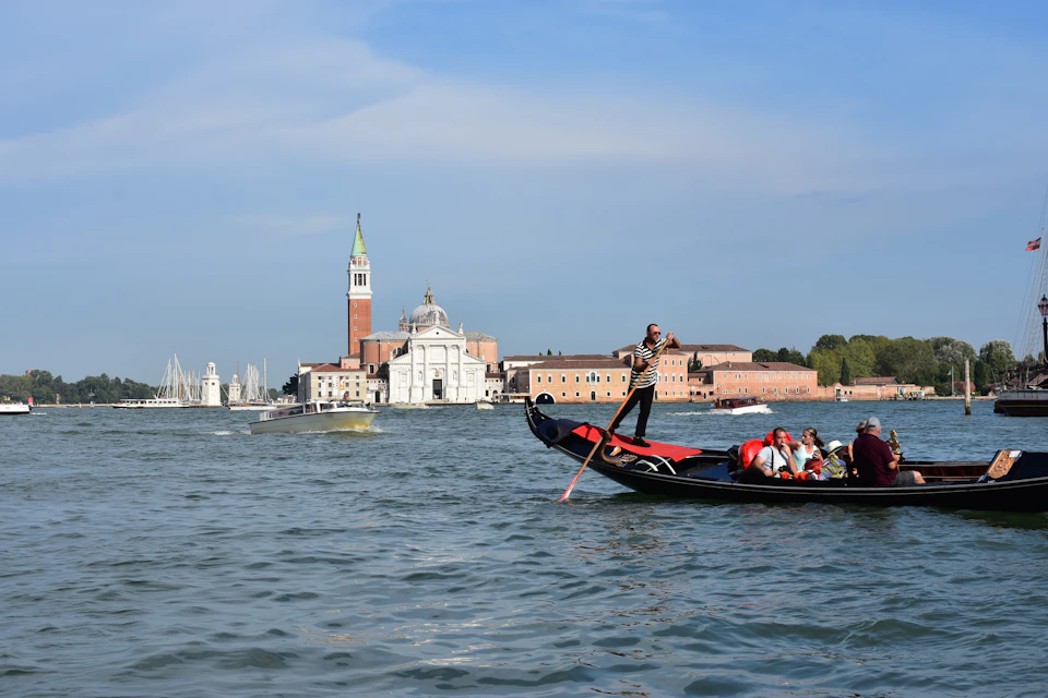 A gondolier rows a gondola with passengers on a canal in front of historic buildings, including a tall tower and church.