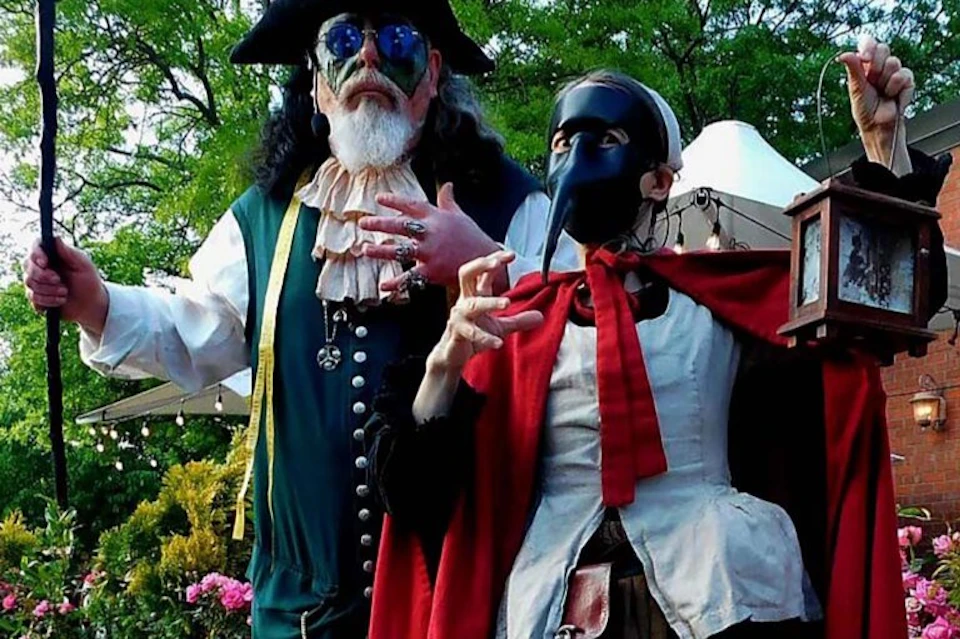 Two people in elaborate costumes, one in a green outfit with a hat and sunglasses, the other in a mask and red cape, outdoors.