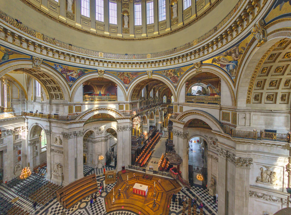Interior of a large, ornate cathedral with columns, arches, and detailed murals. Empty chairs are arranged in rows, and the floor has a checkerboard pattern.