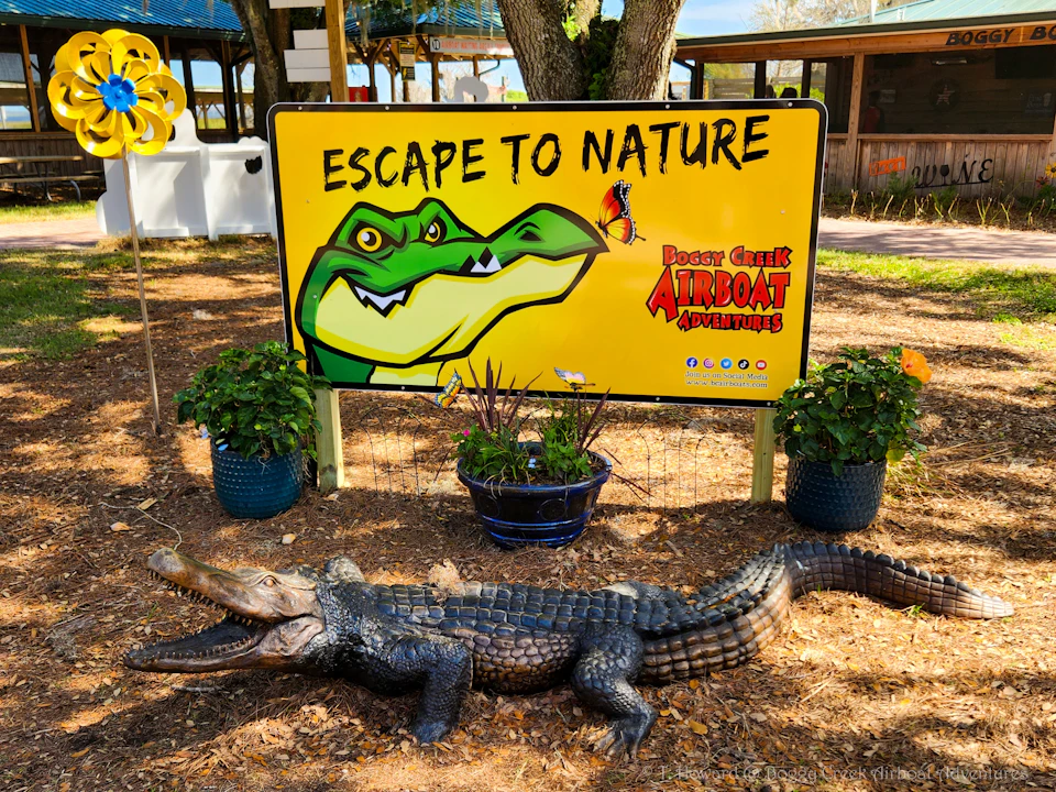 A colorful sign with an alligator graphic and the text "Escape to Nature" and "Boggy Creek Airboat Adventures," surrounded by potted plants.