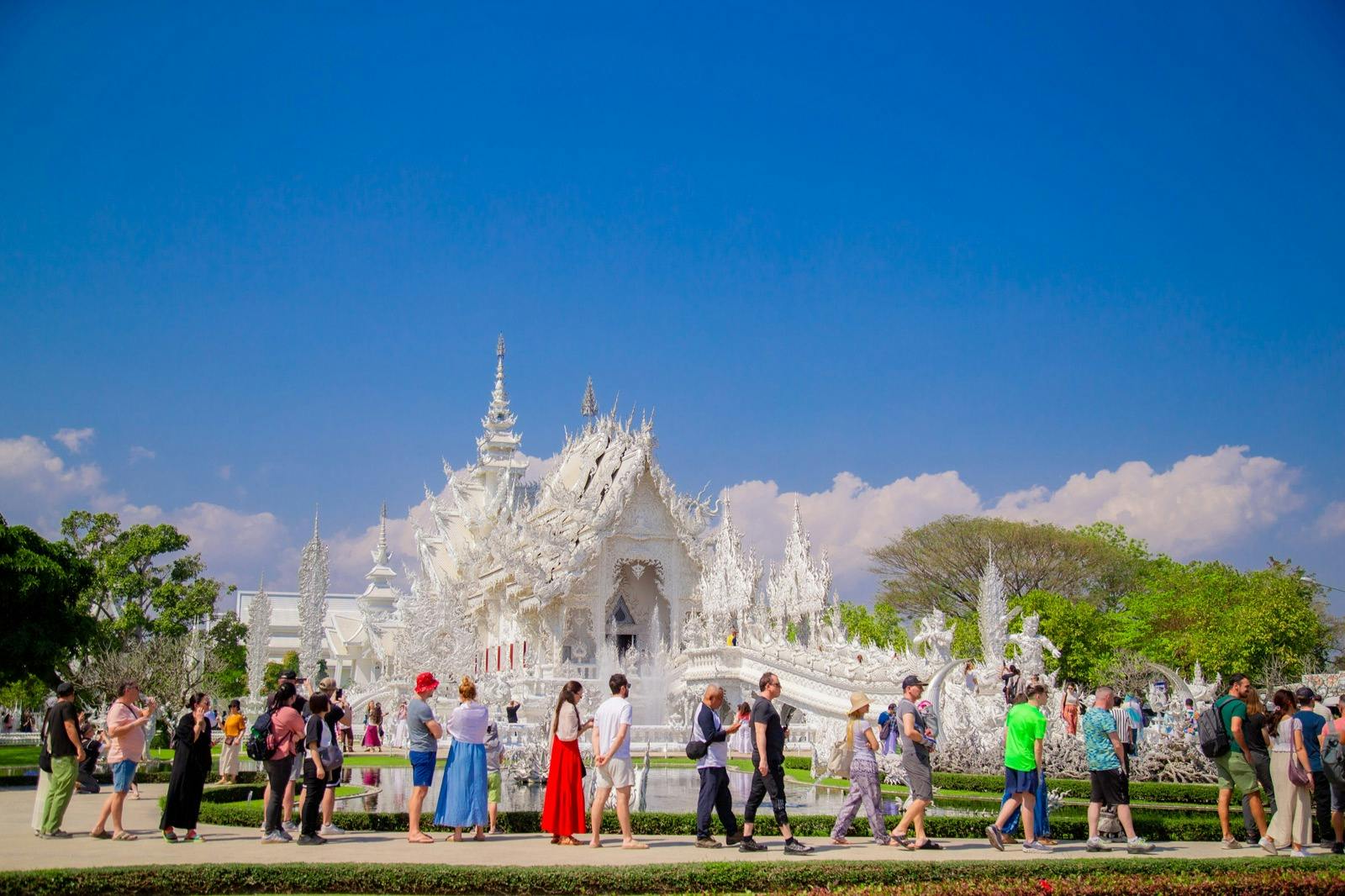 Menschen, die vor einem verschnörkelten weißen Tempel mit komplizierten Details unter einem klaren blauen Himmel spazieren gehen.