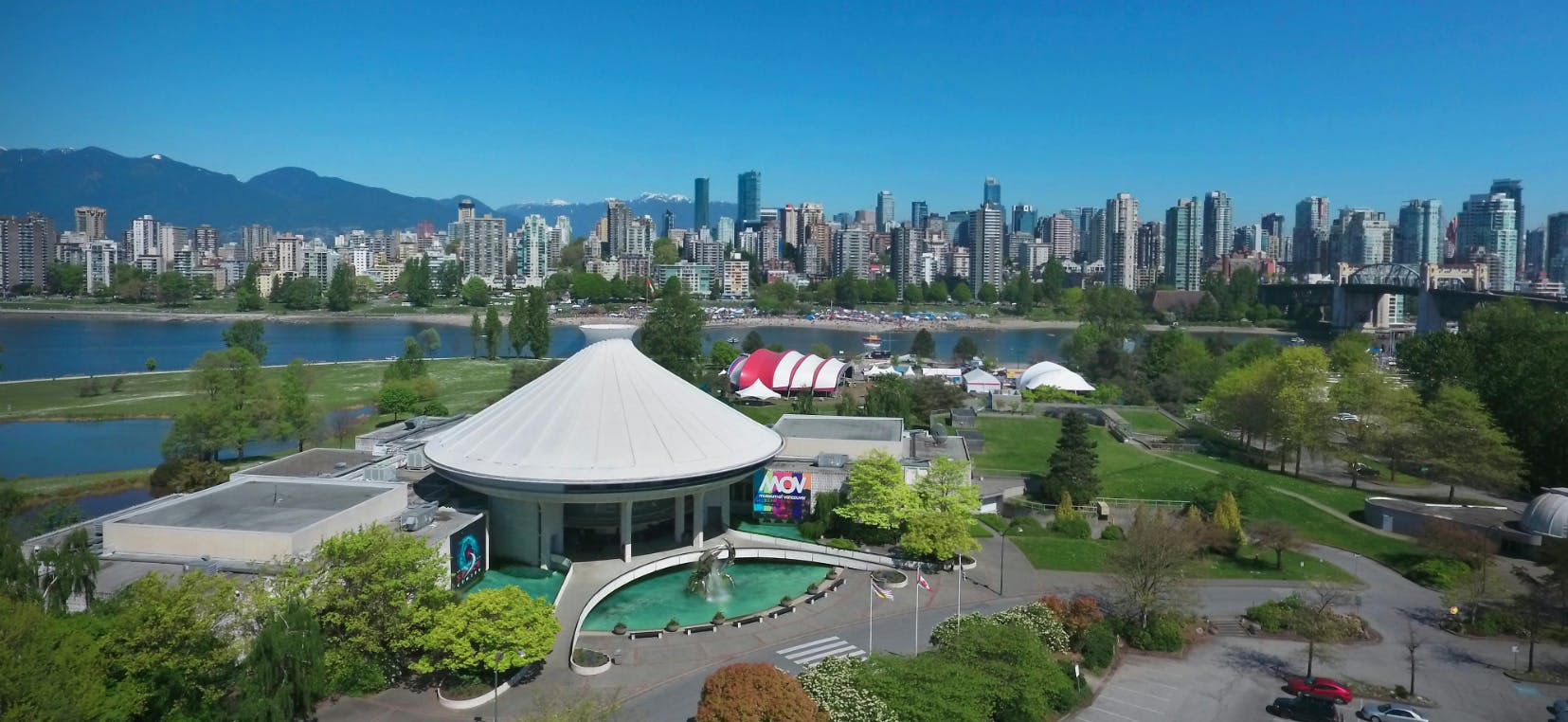 Un paisatge urbà amb un edifici abovedat al primer pla, un riu, espais verds i un skyline d&#39;edificis alts sota un cel blau clar.