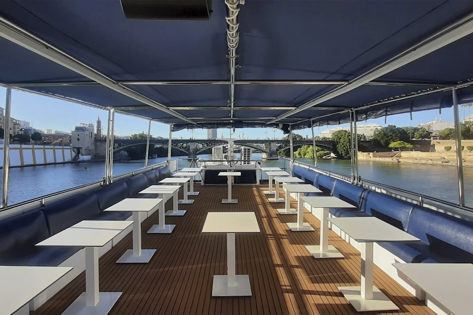 An empty boat deck with rows of white tables, blue seating, and an overhead canopy, floating on a river with a bridge in the background.