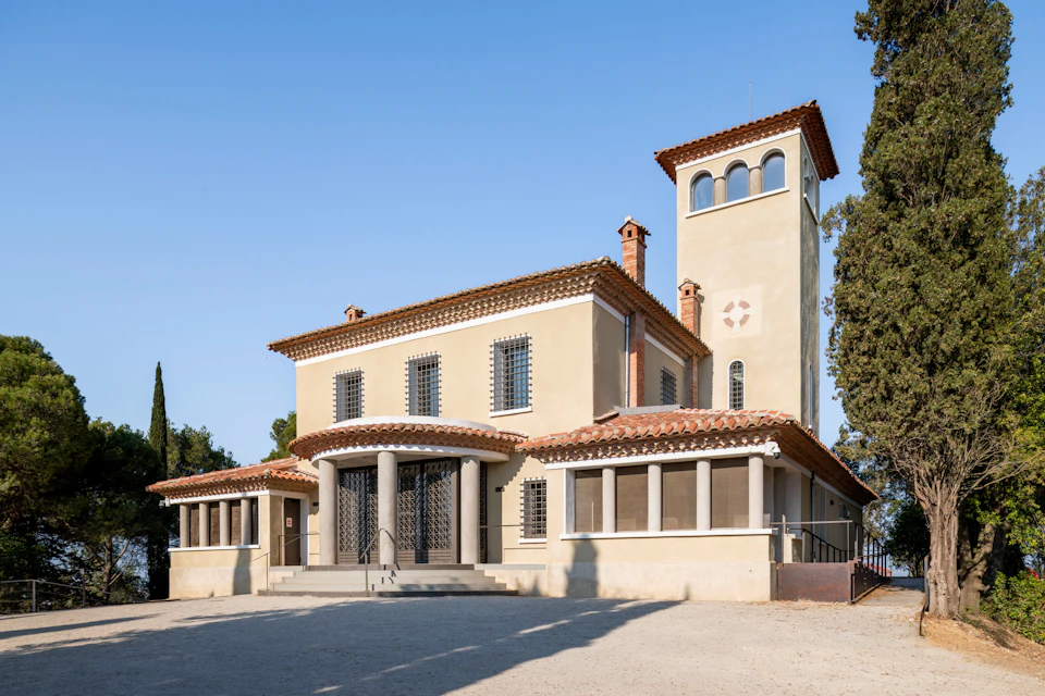 A beige Mediterranean-style building with a tower, red-tiled roof, arched windows, and a columned entrance under a clear sky.