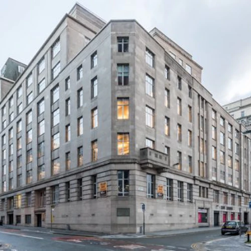 Urbana gathörn med beige byggnader i flera våningar, parkerade bilar och några gående fotgängare under en molnig himmel.