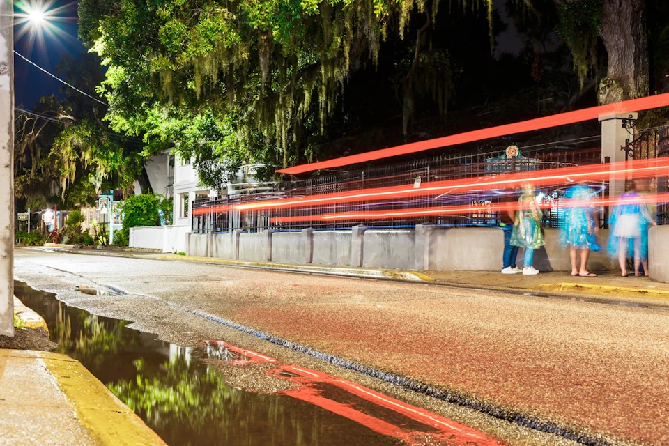 A nighttime urban street scene with light trails from a vehicle, a person walking, and trees with hanging moss in the background.