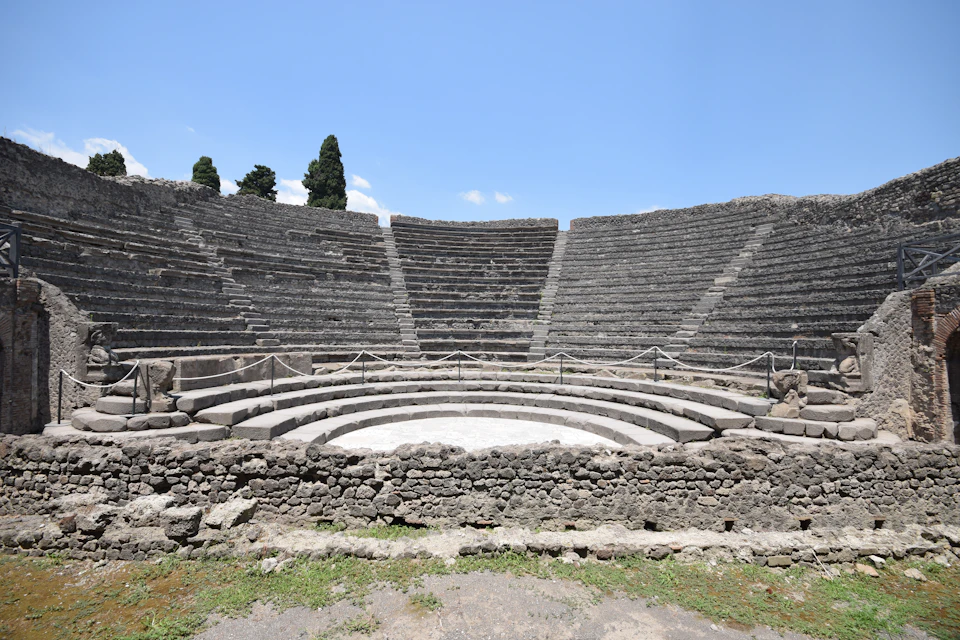 Ancient stone amphitheater with tiered seating under a clear blue sky, surrounded by a few tall trees.