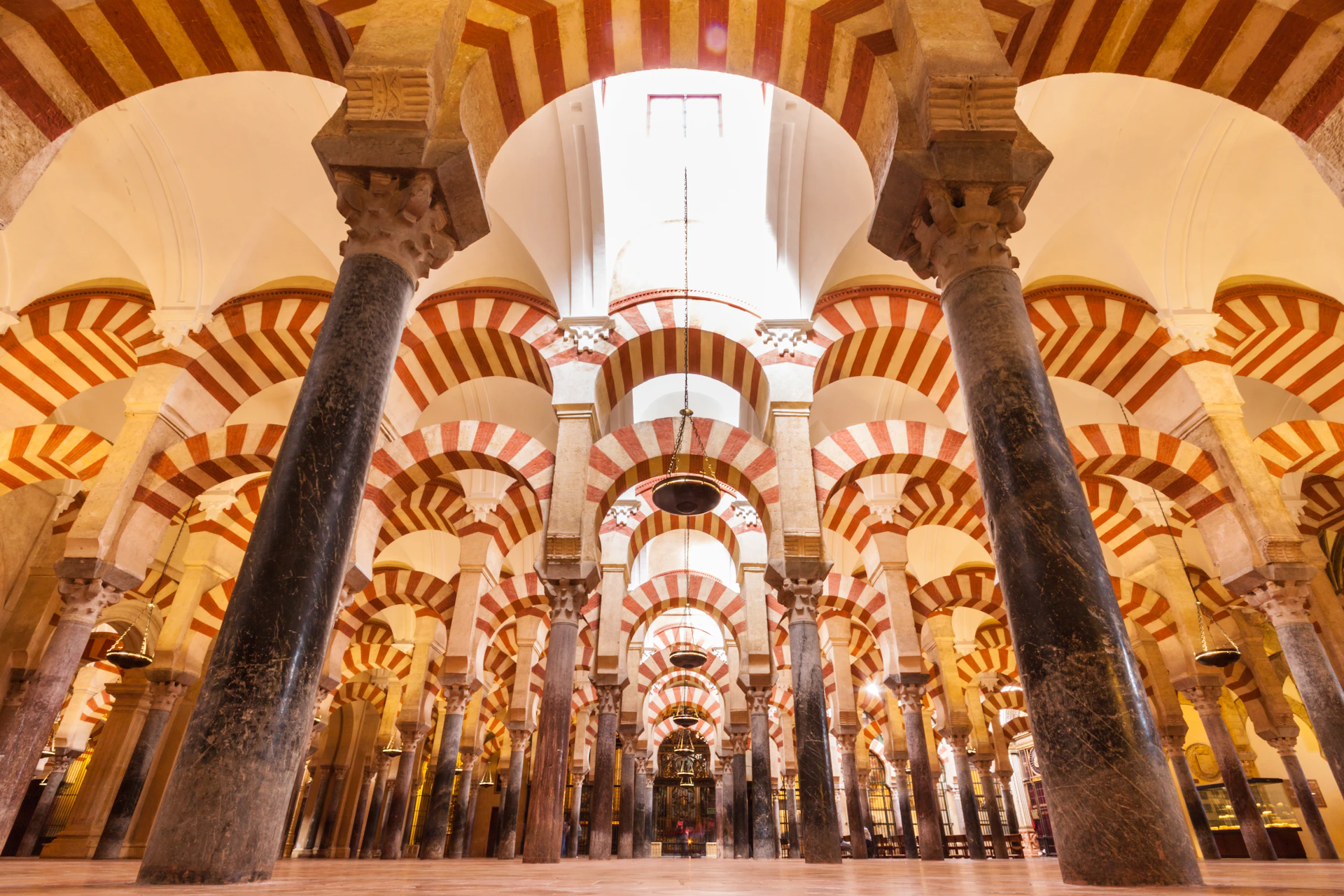 Grand hall with tall, black columns topped with alternating red and white striped arches, illuminated by natural light from above.