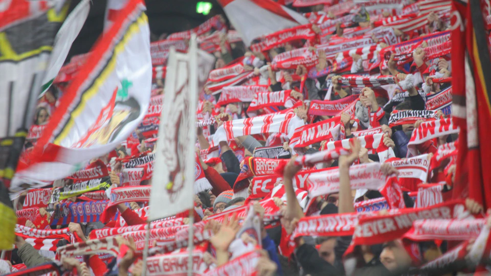 A large crowd of sports fans holding up red and white scarves with team logos and text in a stadium, displaying excitement.