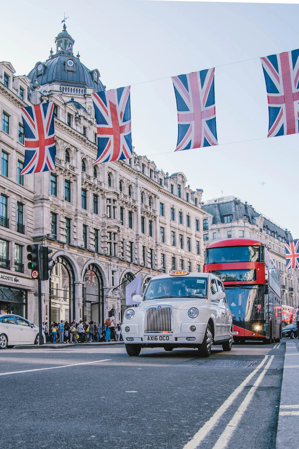 A white taxi and a red double-decker bus drive under Union Jack flags on a busy street in front of historic buildings.