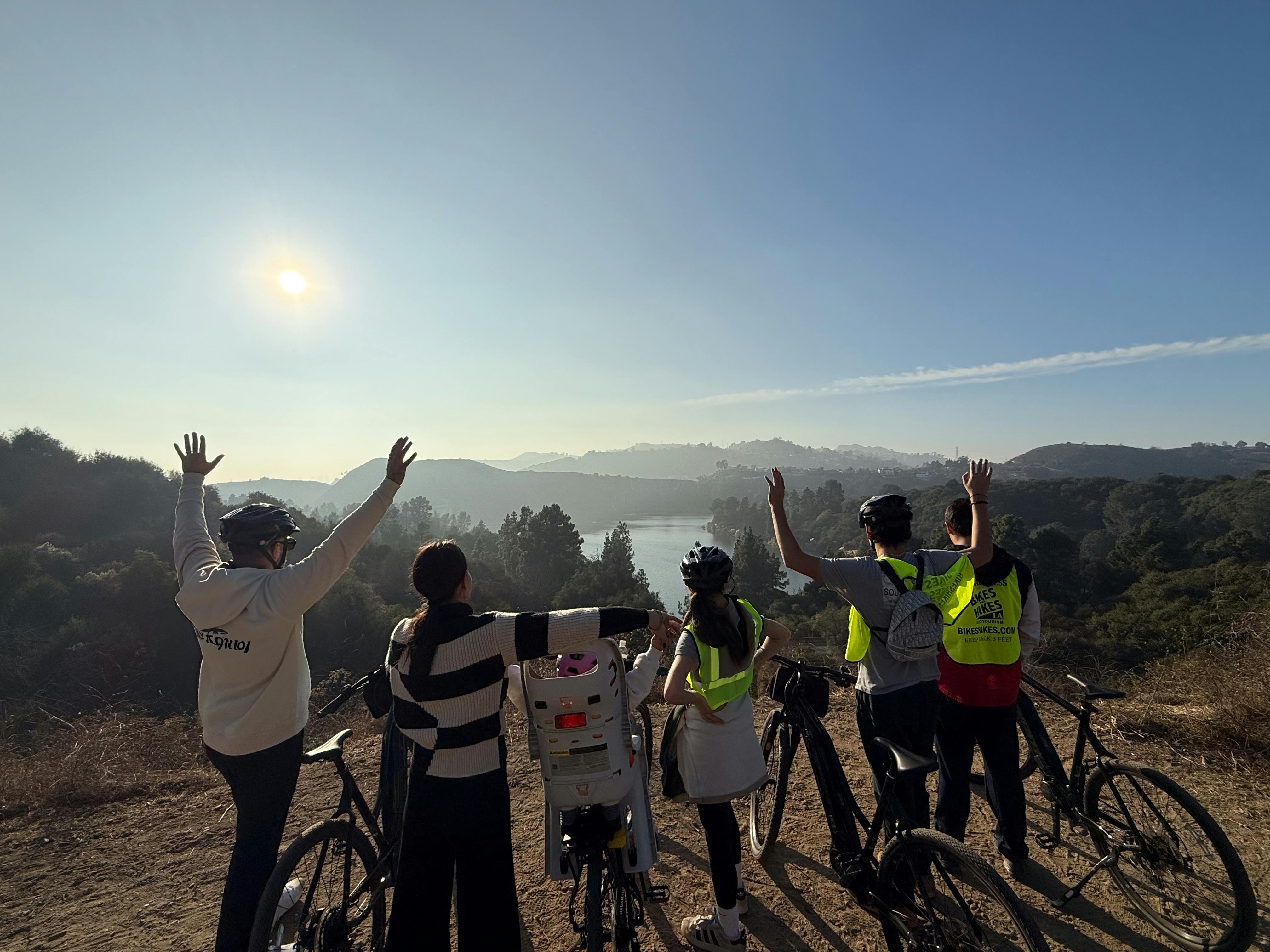 Five people in helmets stand with bicycles, raising hands, overlooking a scenic landscape with hills and a lake under a sunny sky.