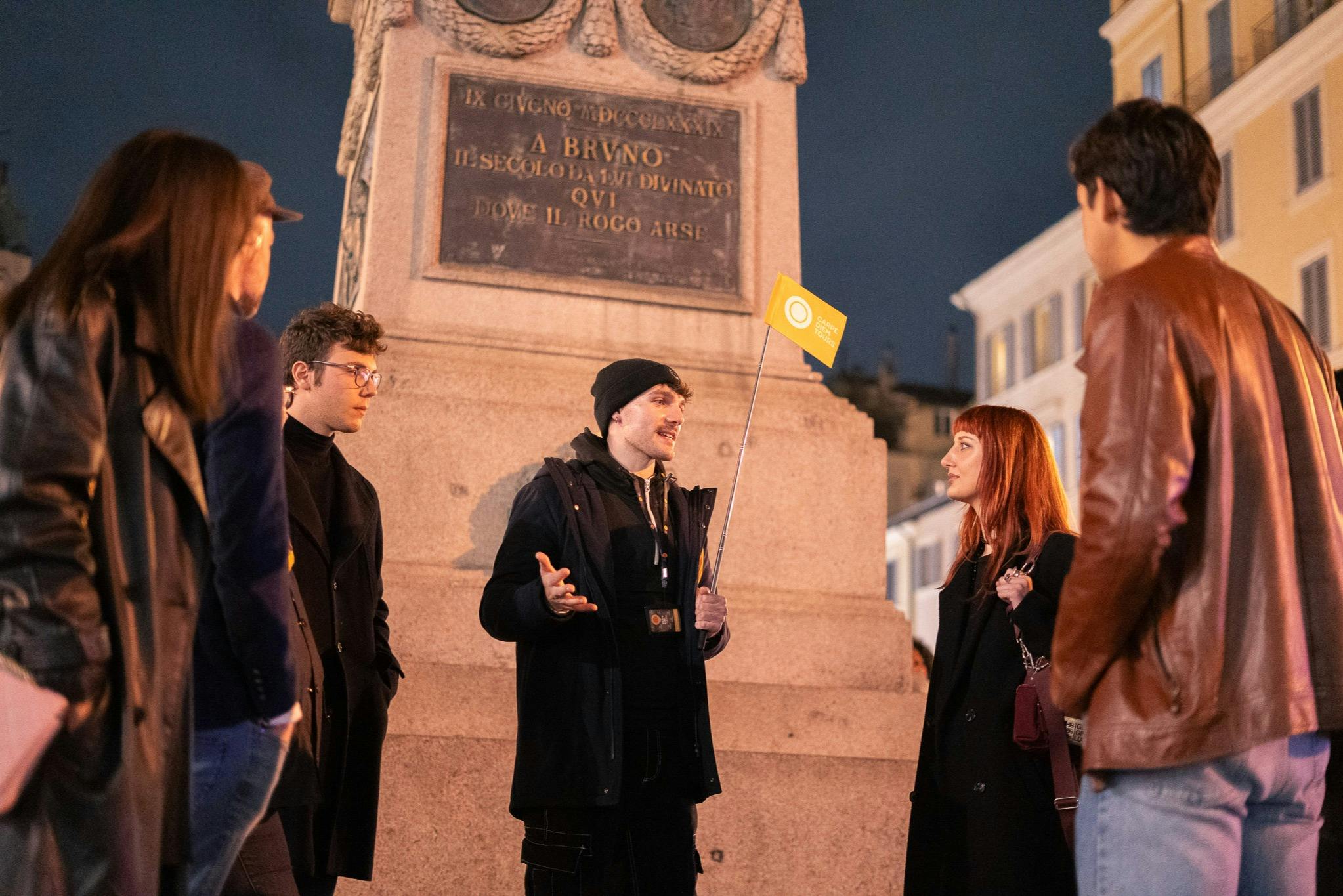 A group of people standing near a monument at night, one holding a small yellow flag.