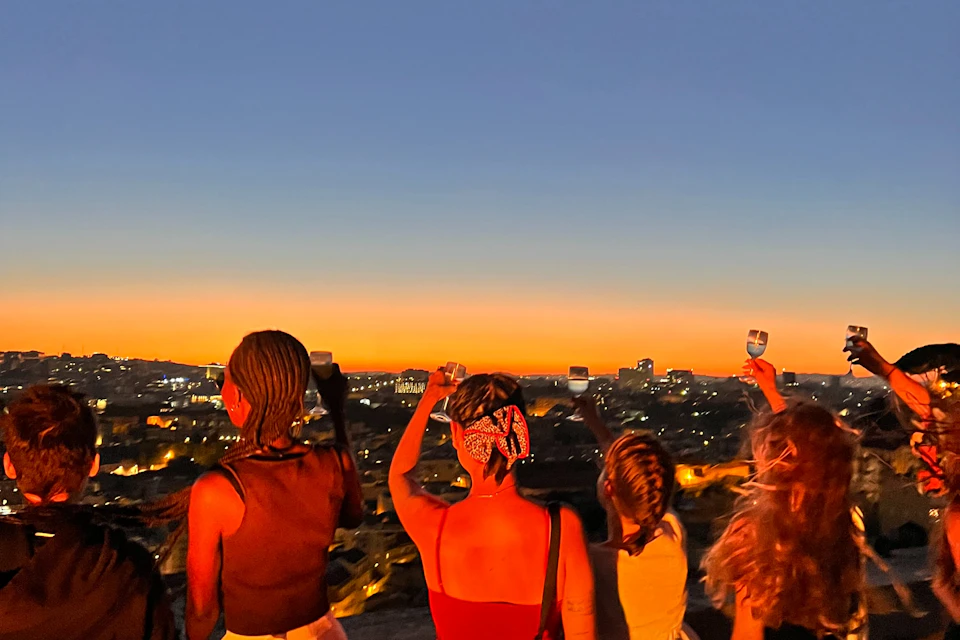 Four people with raised wine glasses overlook a cityscape at sunset, with a gradient sky transitioning from orange to deep blue.