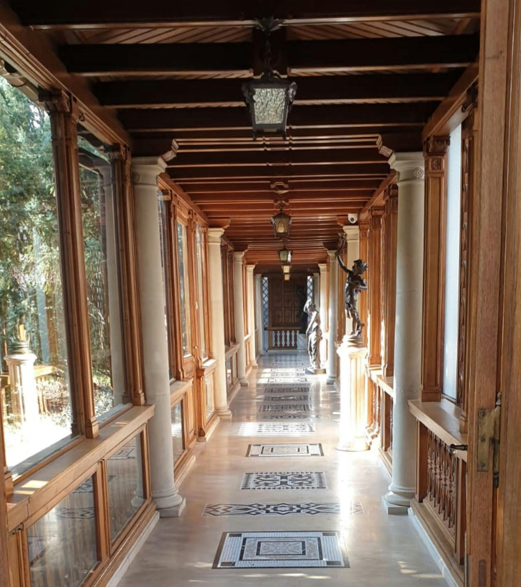Un passadís de fusta amb finestres de vidre a l'esquerra, estàtues, columnes i rajoles estampades que condueixen a una porta.