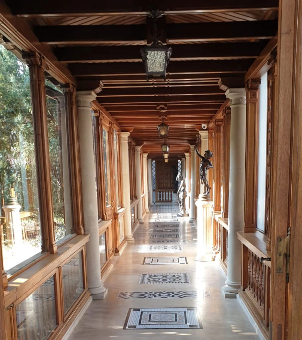 A wooden hallway with glass windows on the left, statues, columns, and patterned floor tiles, leading to a door.