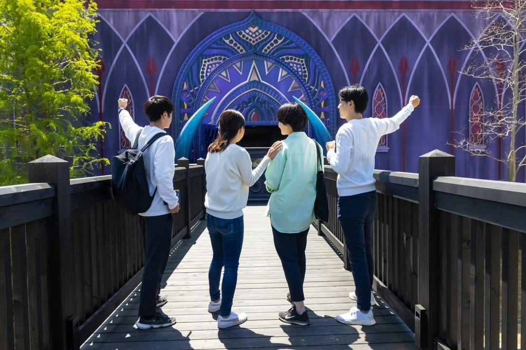 Three people stand on a wooden bridge in front of a colorful, patterned arched building facade.