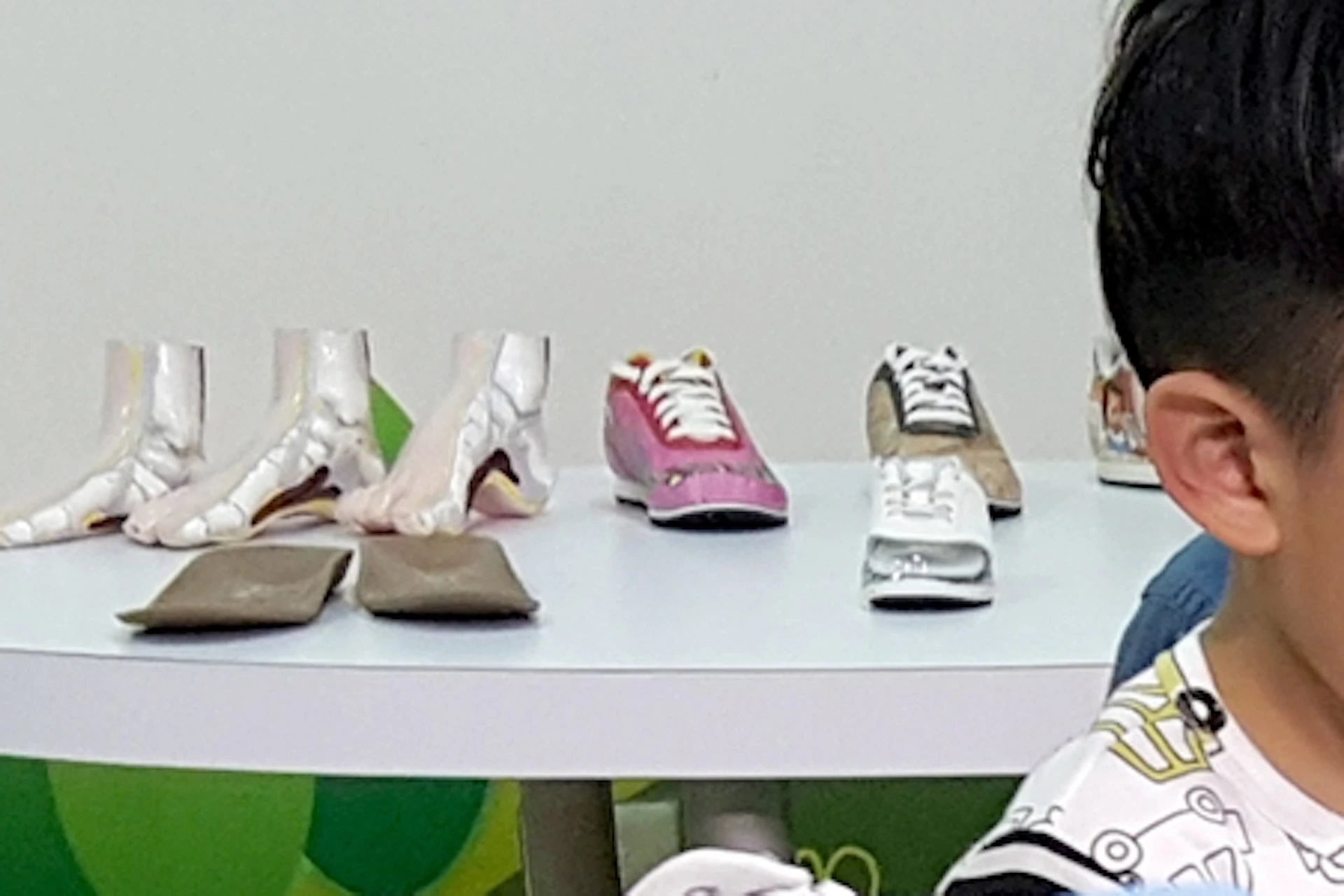 A woman, child, and man sit at a table with papers and a shoe design activity, with shoes displayed on a table in the background.