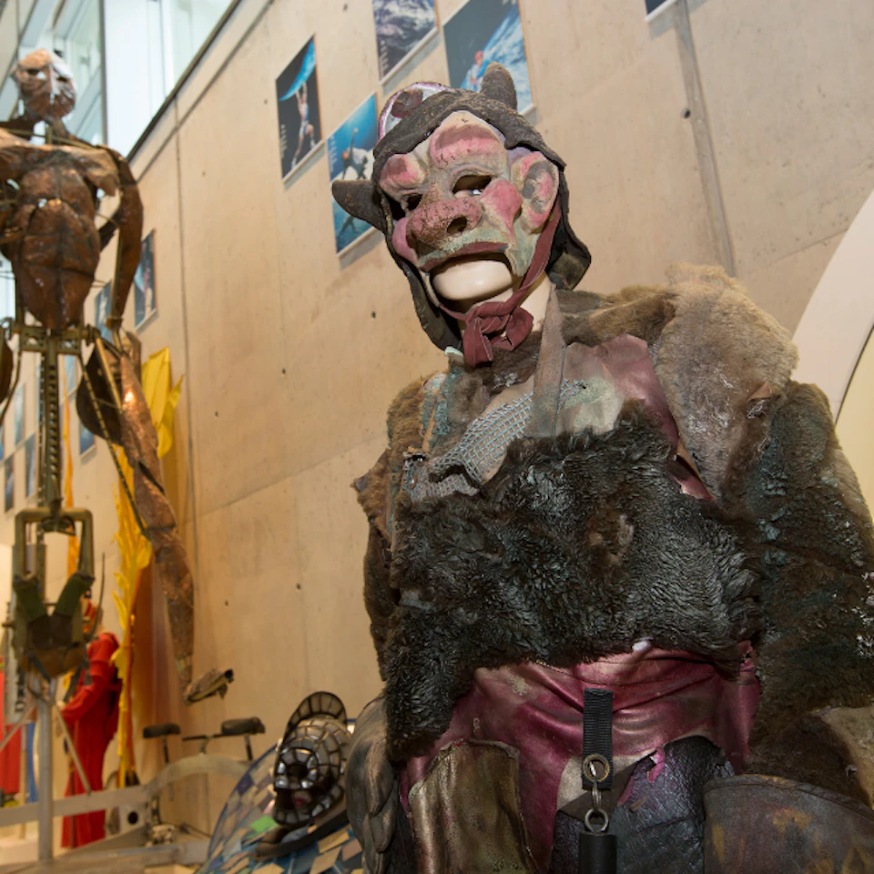 Person wearing a detailed, monster-like costume, including a mask and fur. Background includes sculptures and a concrete wall.