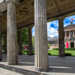 Rad av stenpelare som ramar in en vy av en neoklassisk byggnad med rosa banderoller och en staty, placerad mot en vibrerande blå himmel.