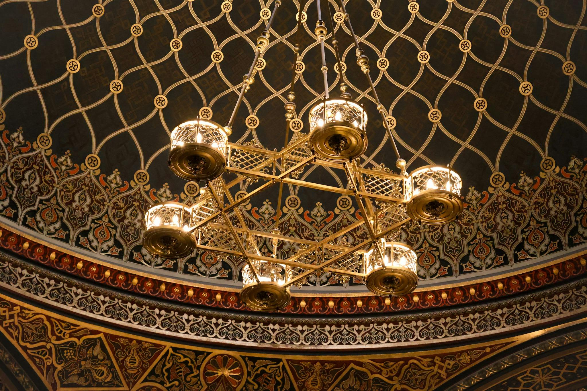 A golden chandelier with intricate designs hangs from a dome ceiling with elaborate geometric and floral patterns.