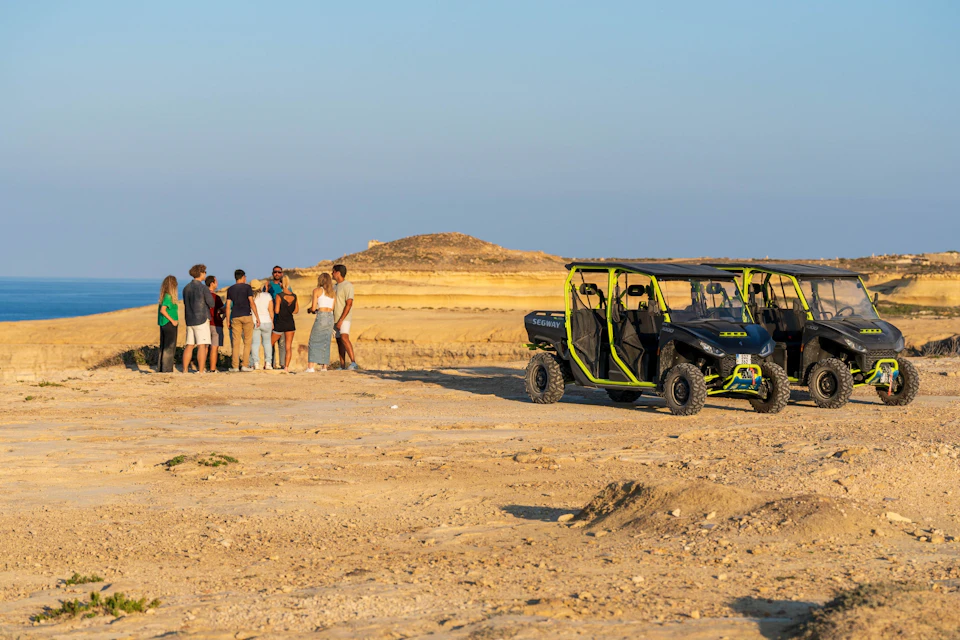 A group of people stands near two off-road vehicles on a desert landscape with a hill in the background under a clear sky.